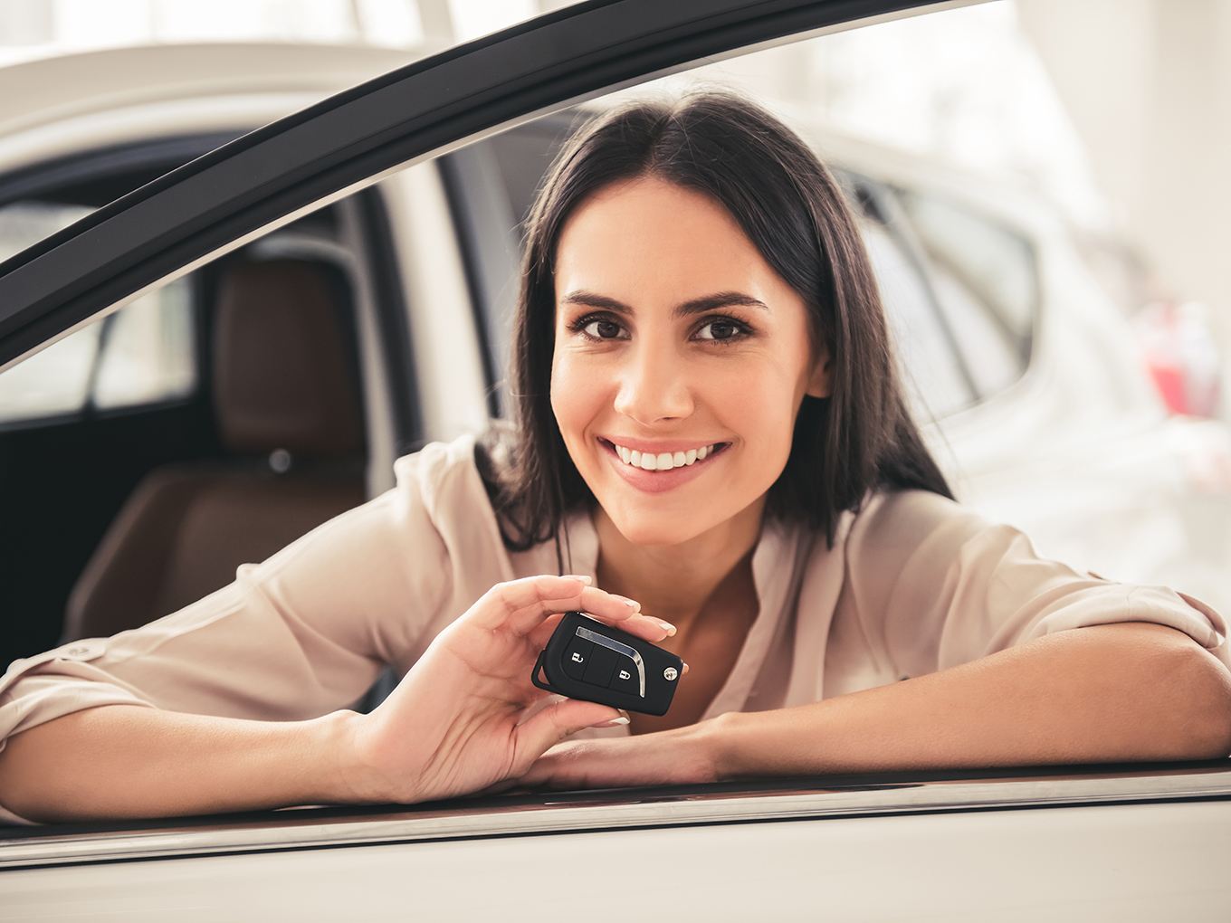A happy young woman sits behind the wheel of a car, holding car keys and smiling.