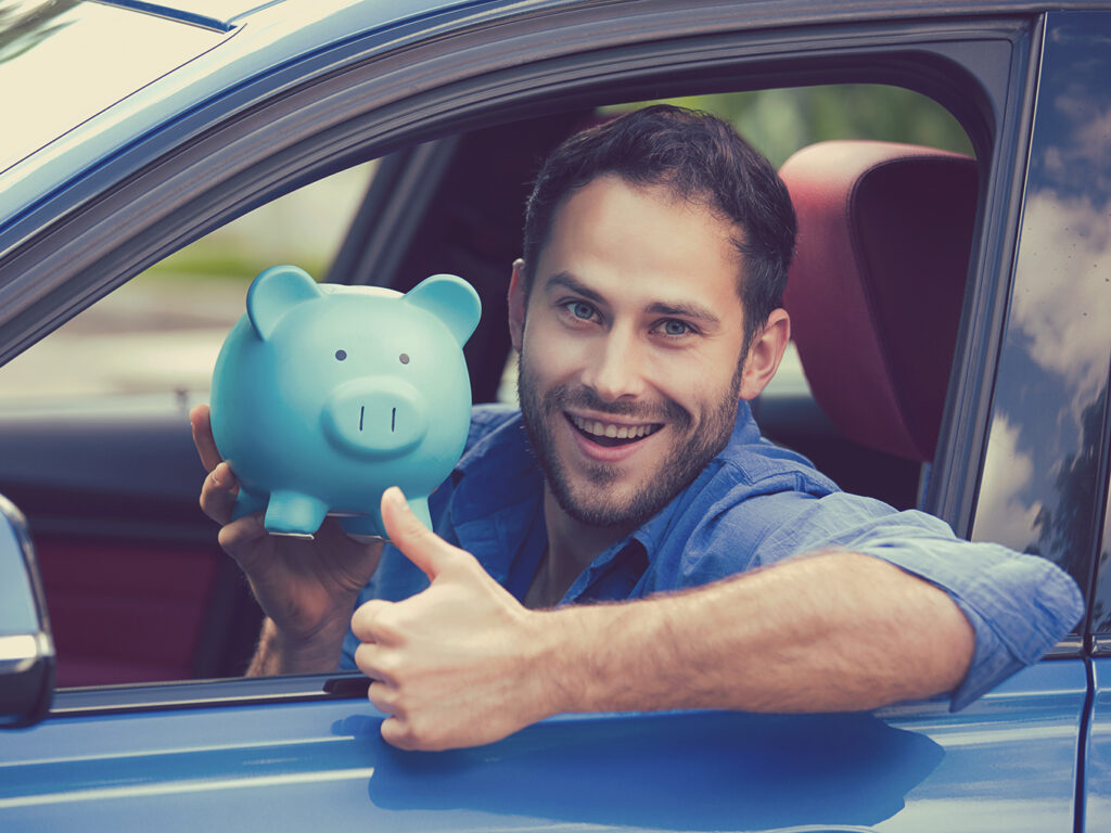 A man sitting inside a new car holding a piggy bank and showing thumbs up, symbolizing savings through car refinance loans.