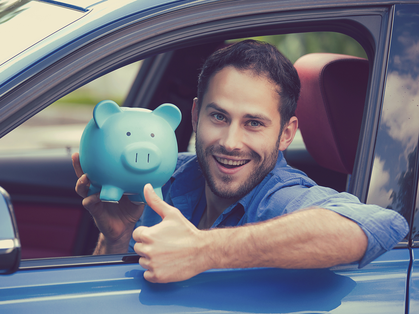 A man sitting inside a new car holding a piggy bank and showing thumbs up, symbolizing savings through car refinance loans.