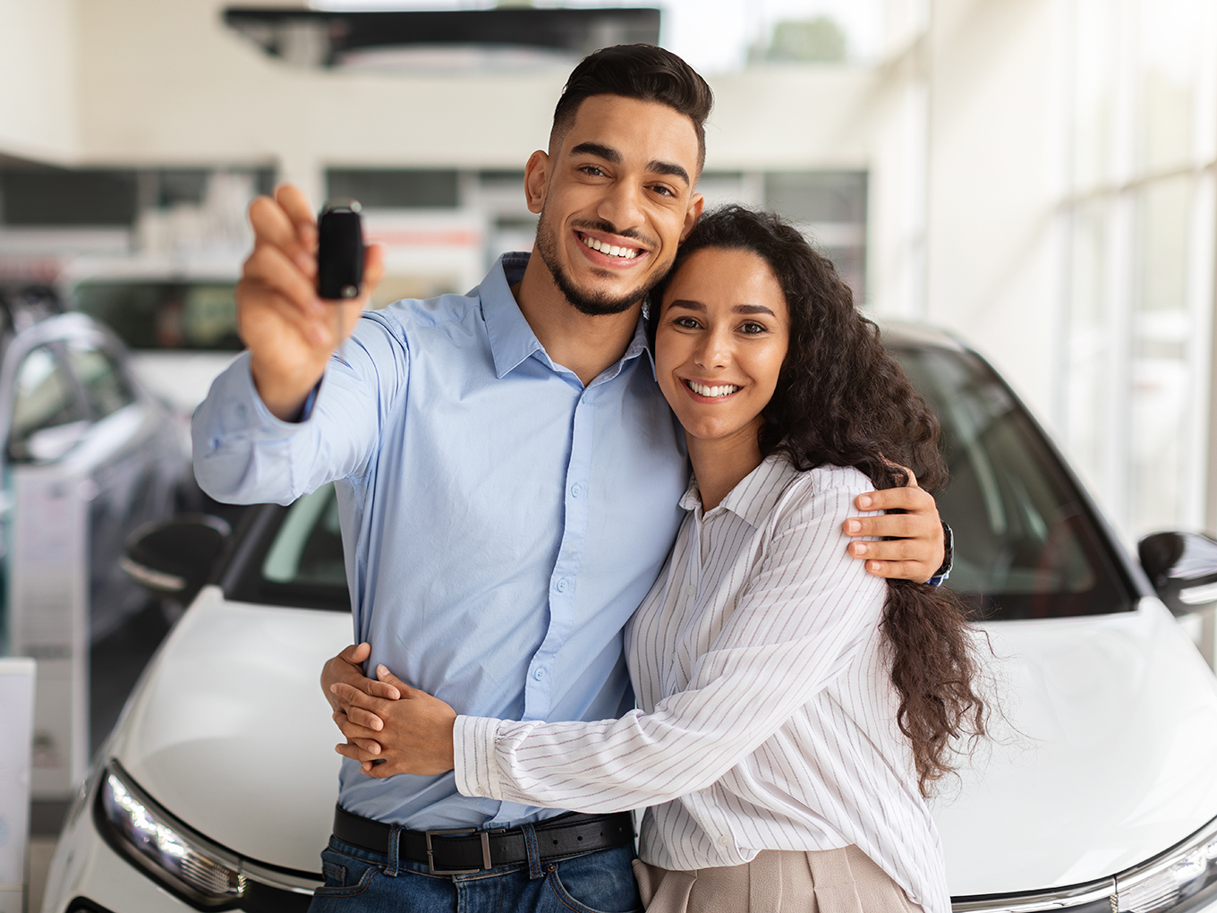 A happy Middle Eastern couple holding up a car key and hugging in front of their new vehicle at a dealer showroom, after using Approval Heroes for a hassle-free car-buying experience.