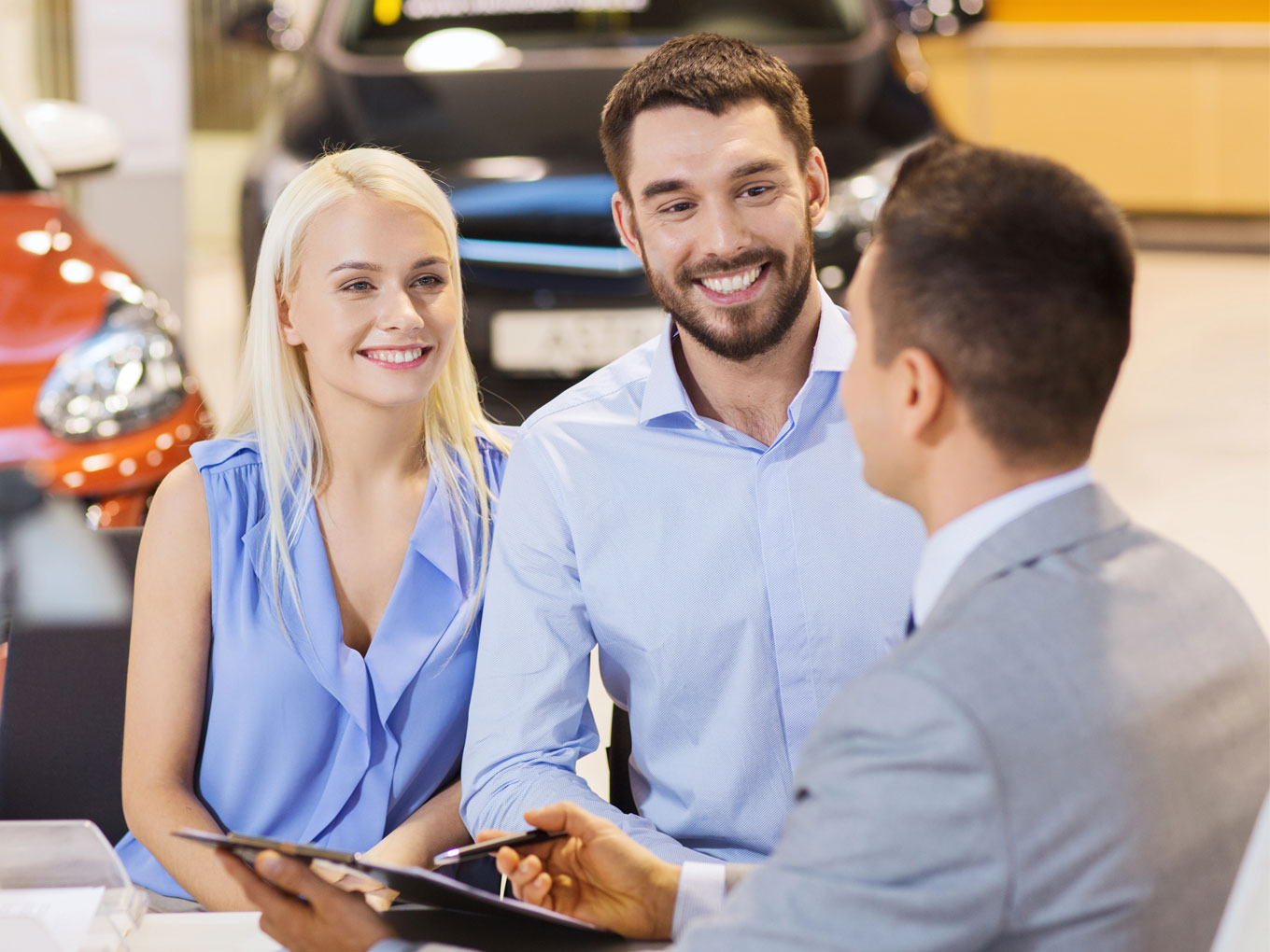 A smiling couple sitting across from a car dealer in a dealership, discussing car purchasing options.