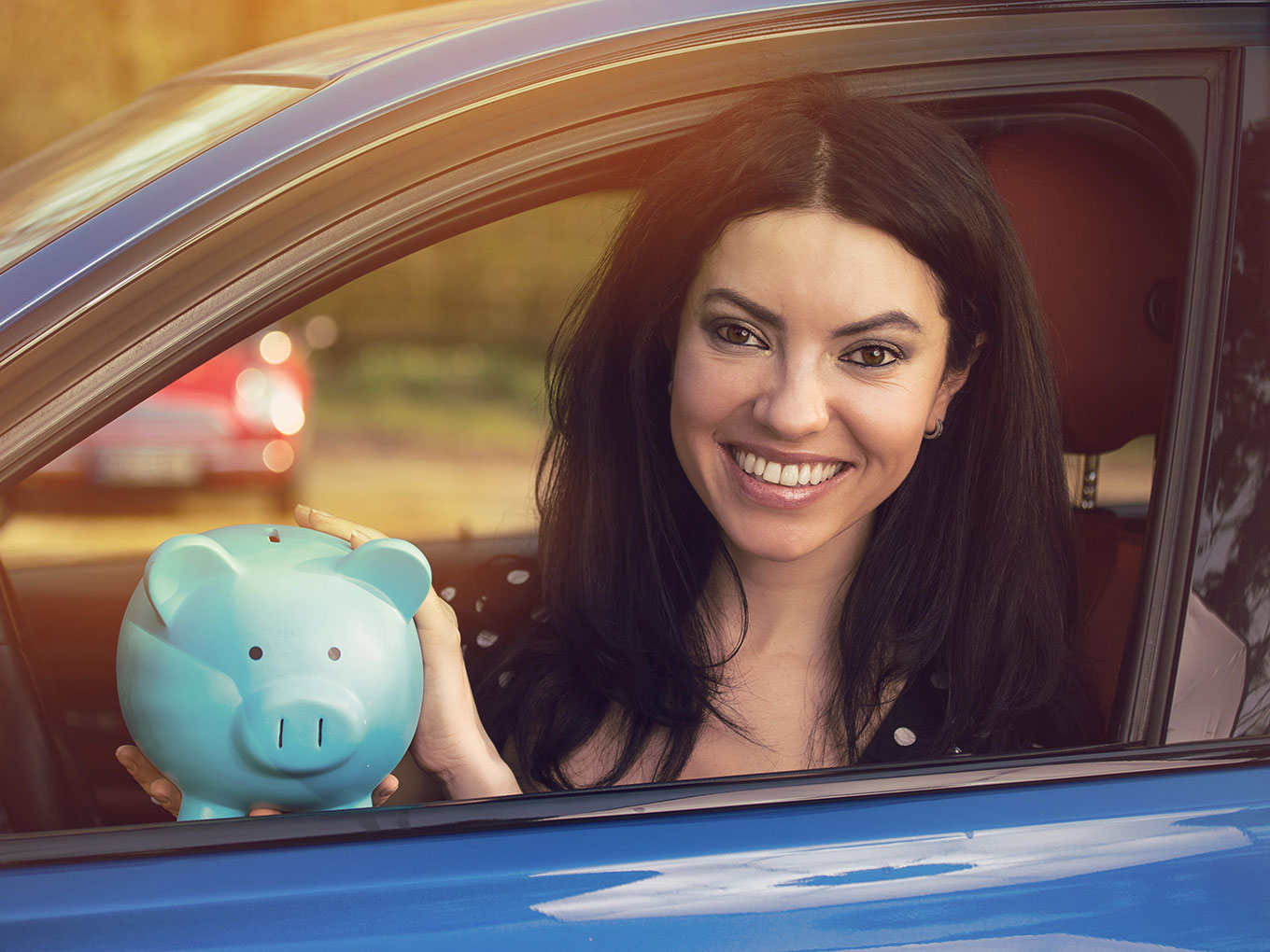 A smiling woman sitting in her car, holding a blue piggy bank, symbolizing savings on auto loans