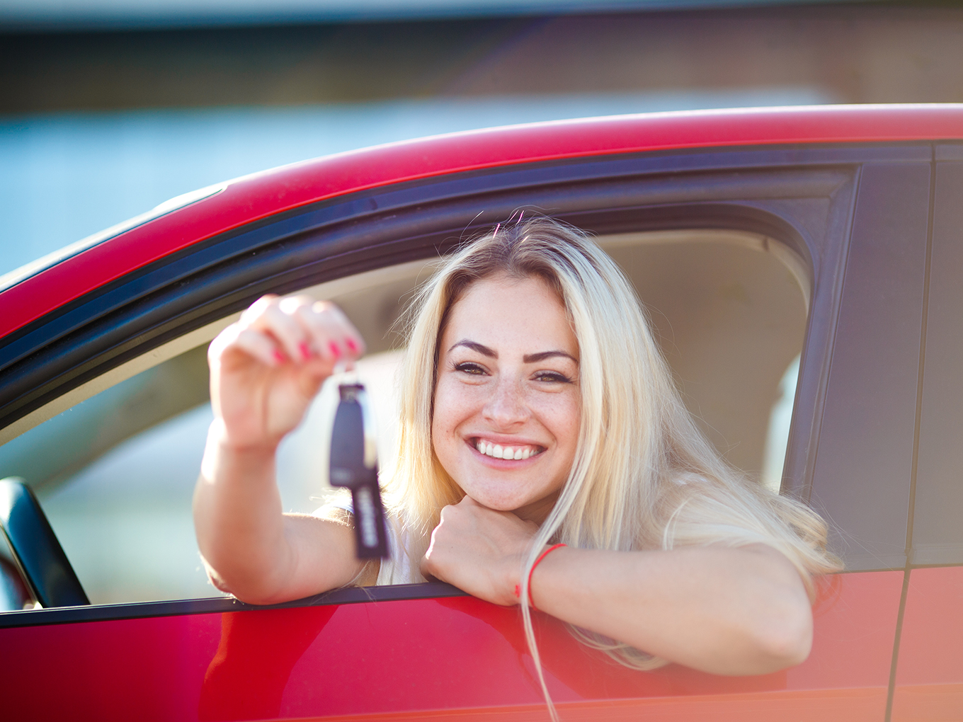Happy woman with blonde hair sitting in her new red car, holding car keys with the car window open