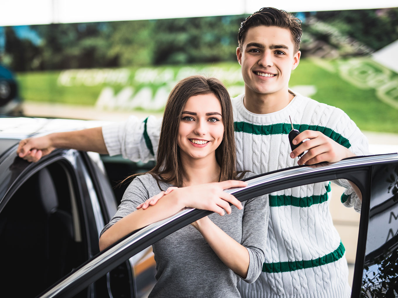 A happy young couple leaning on their new car, smiling and looking at the camera, with the man holding the car keys